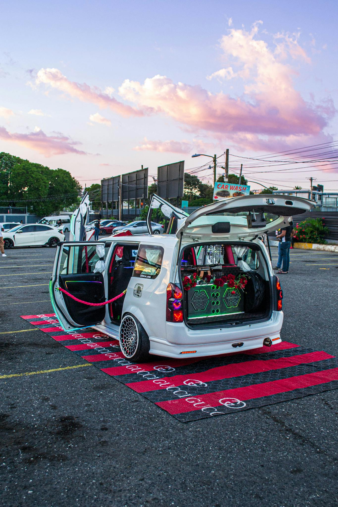 A custom white car with open doors and neon lights in a parking lot at sunset.
